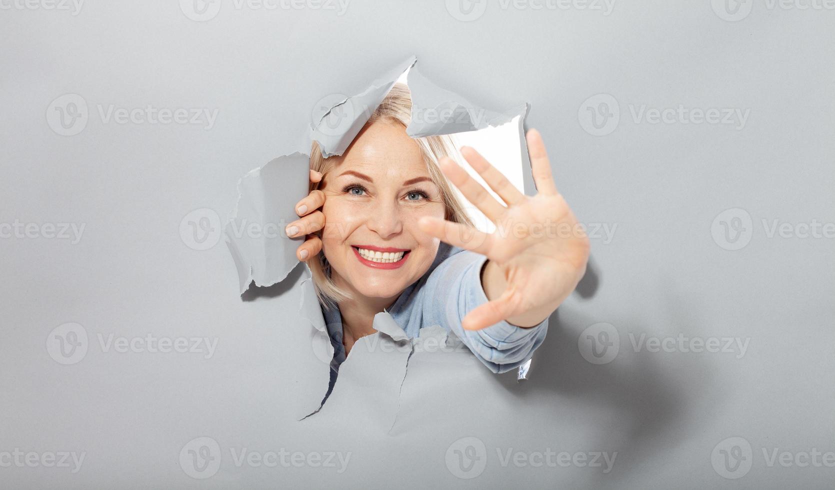 atractiva y alegre mujer de mediana edad que agita un gesto de saludo con la mano, muestra que todo está bien, todo está en orden. agujero rasgado en papel gris. foto
