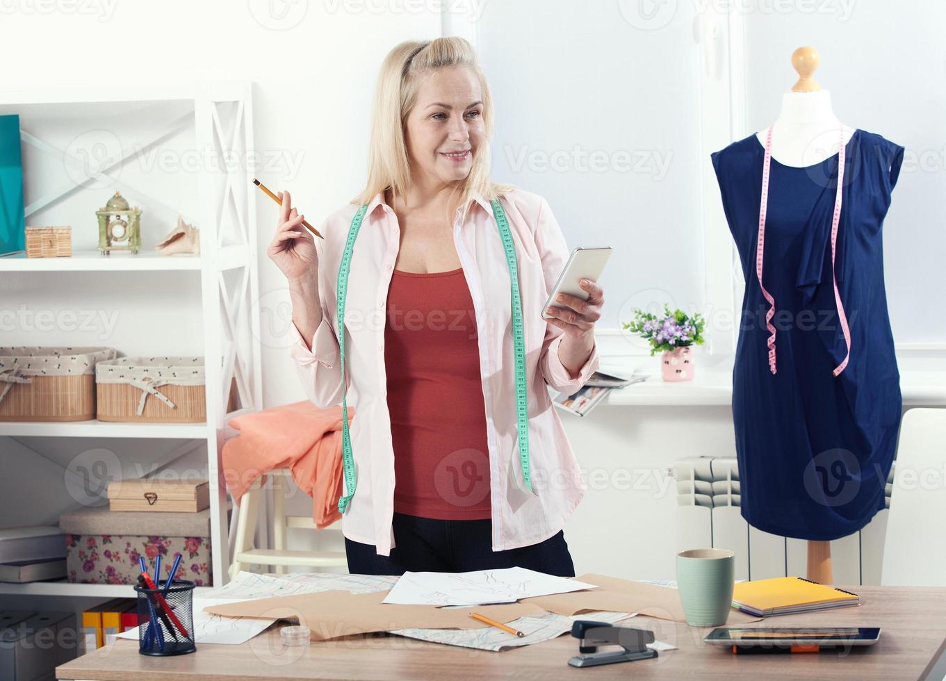 Communication with suppliers by phone, skype. Beautiful European woman tailor talking on the phone, standing near the table in the workshop with clothes hanging in background photo