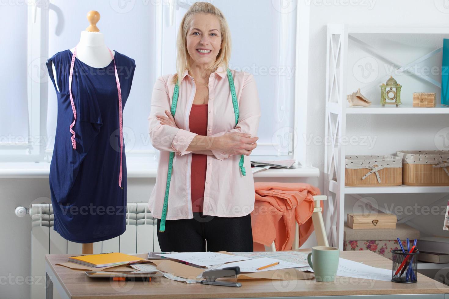 A successful woman designer clothes, tailor in the studio in full growth. Beautiful European woman looking at the camera, standing near the table in the workshop with clothes hanging in the background photo