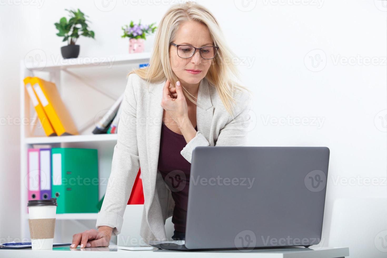 Working on new project. Business woman with glasses working in office with documents. Beautiful middle aged woman looking at laptop with smile while siting in the office. photo