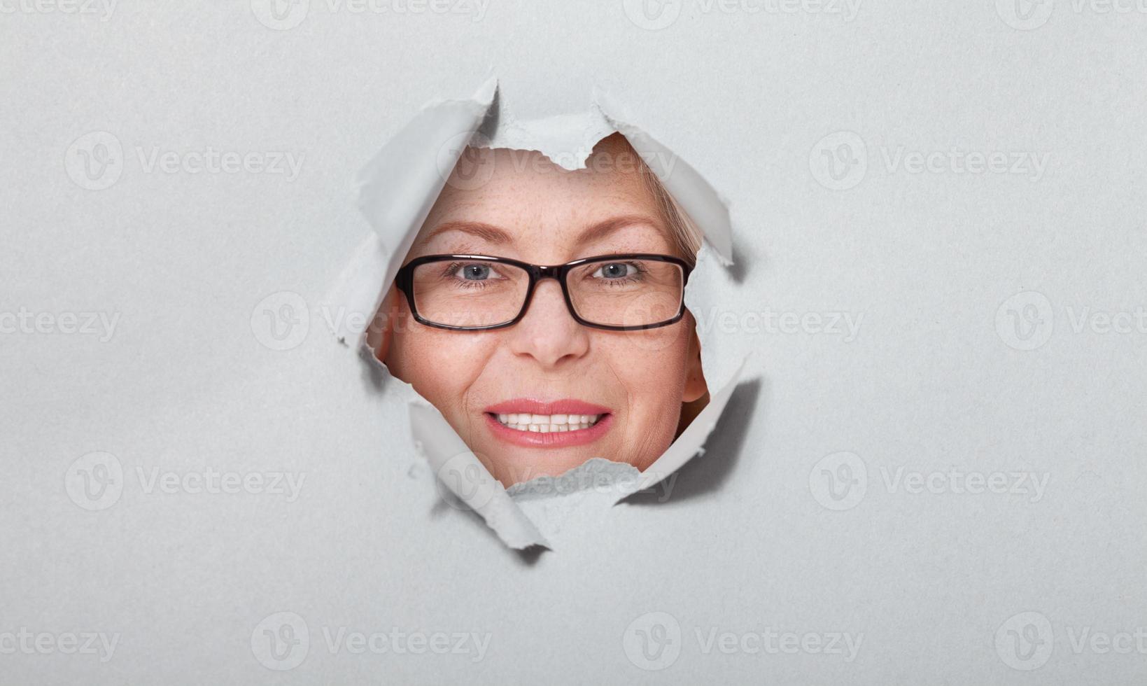 Surprised woman in glasses looking playfully in torn paper hole, has excited cheerful expression, looks through breakthrough of gray background. Wow, great news photo