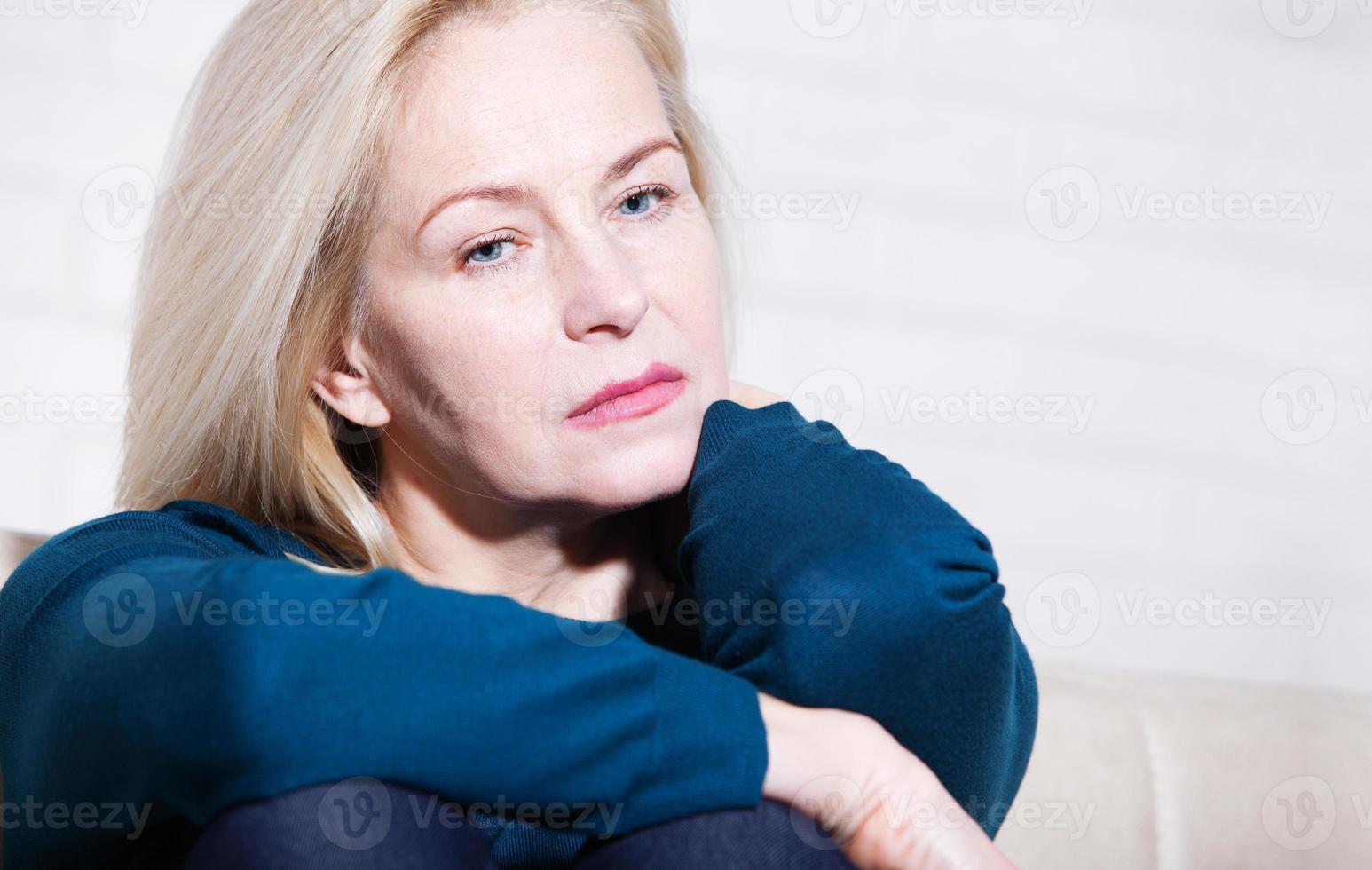 Middle aged woman barefoot sitting on the couch, hugging her head, at home. The concept of sadness and loneliness. photo