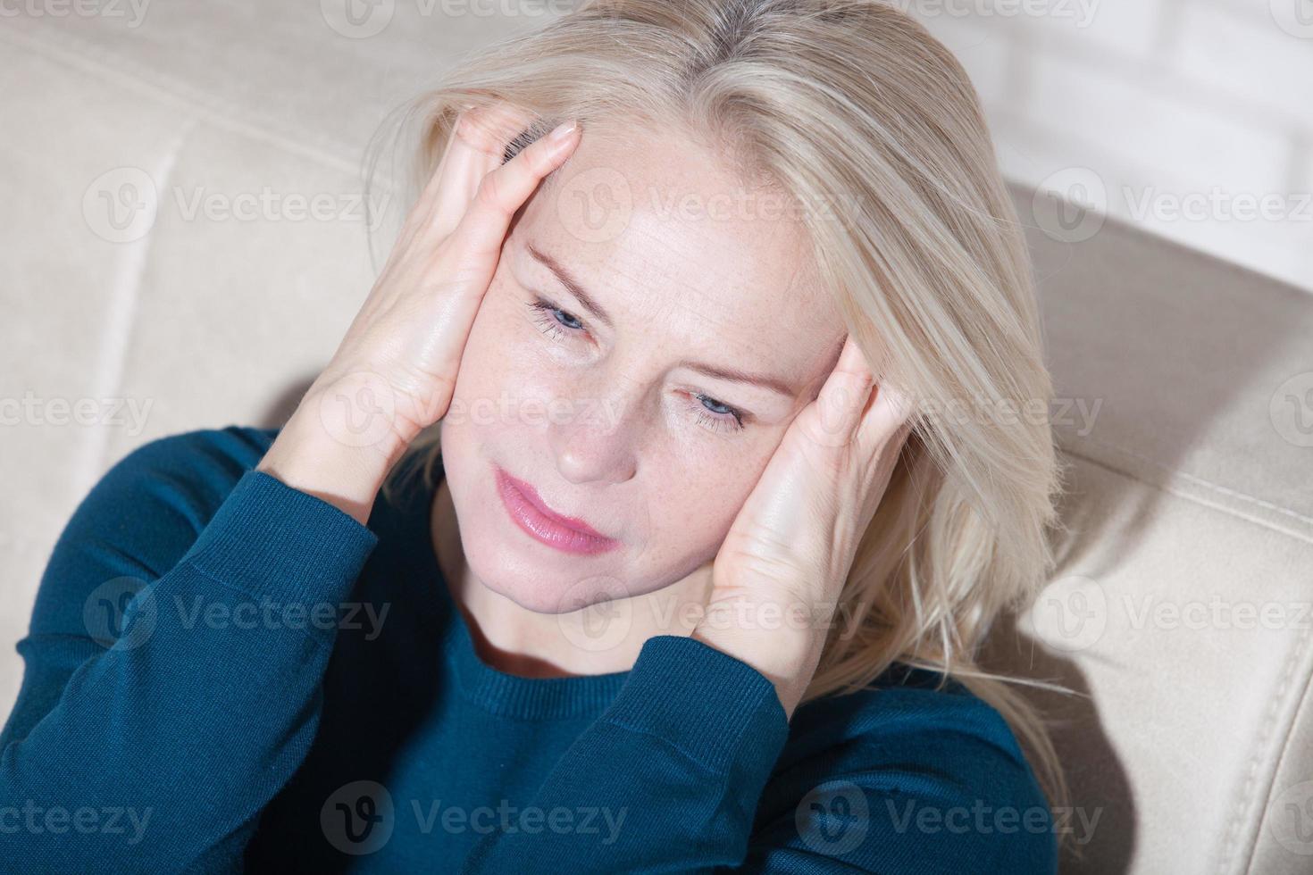 Sad depressed middle aged woman at home sitting on the couch, looking down and touching her forehead, loneliness and pain concept photo