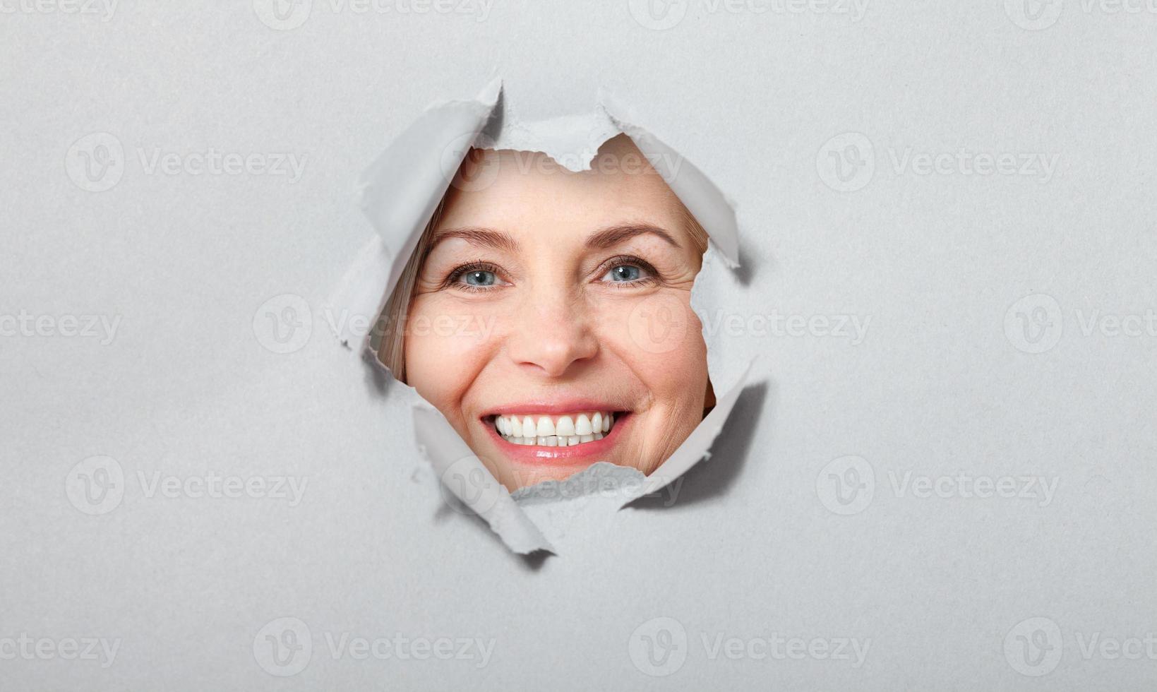 Surprised woman looking playfully in torn paper hole, has excited cheerful expression, looks through breakthrough of gray background. Wow, great news photo