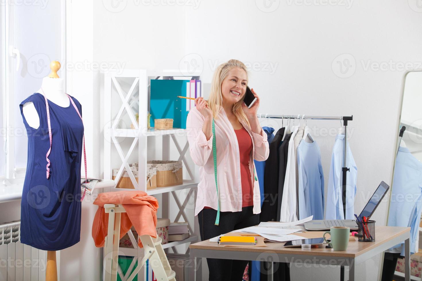 Search for new ideas for business. Beautiful European woman working in the office, standing near the table in the workshop with clothes hanging in the background. Interier design studio. photo