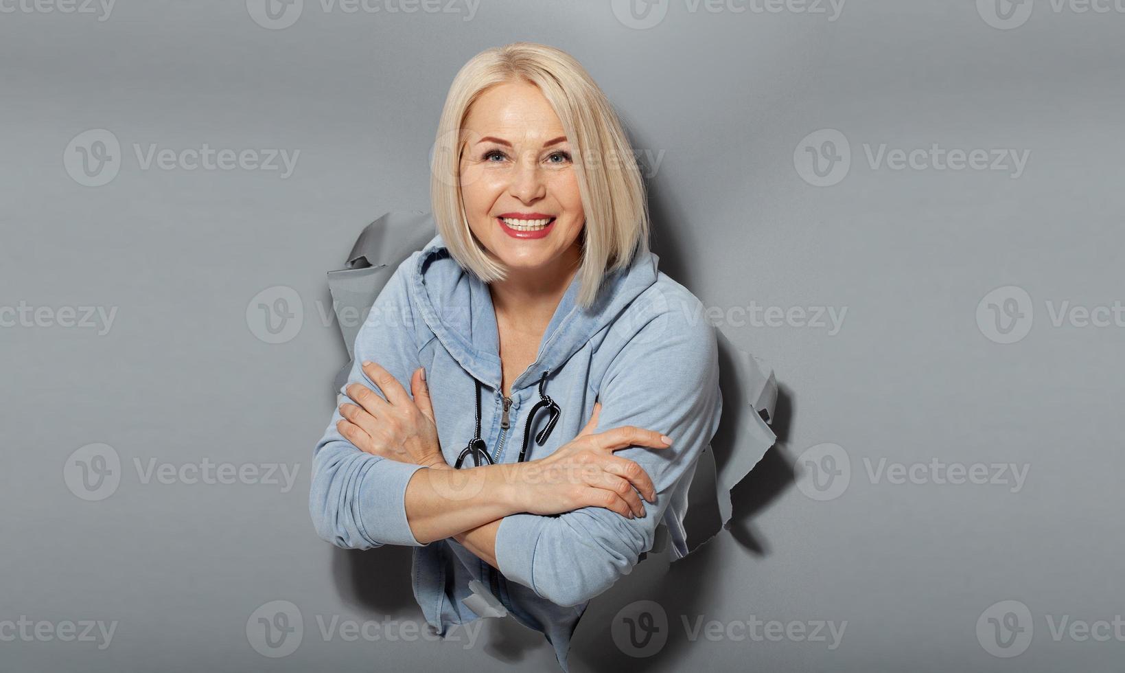 Charismatic joyful woman with blonde hairstyle, looking playfully, wears fashionable hoody, stands in torn paper hole, notices funny pleasant. Gray background. photo