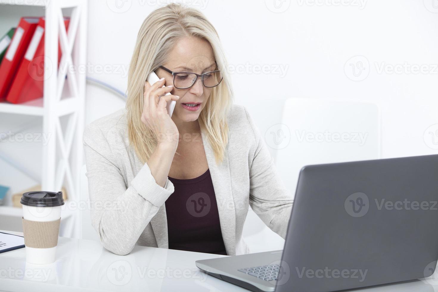 Listening to clients requirements. Beautiful middle aged woman with glasses talking on smart phone and smiling at her working place siting in the office. photo