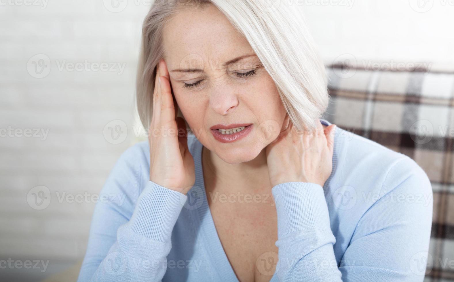 Woman suffering from stress or a headache grimacing in pain as she holds the back of her neck with her other hand to her temple, with copyspace. Concept photo with indicating location of pain.