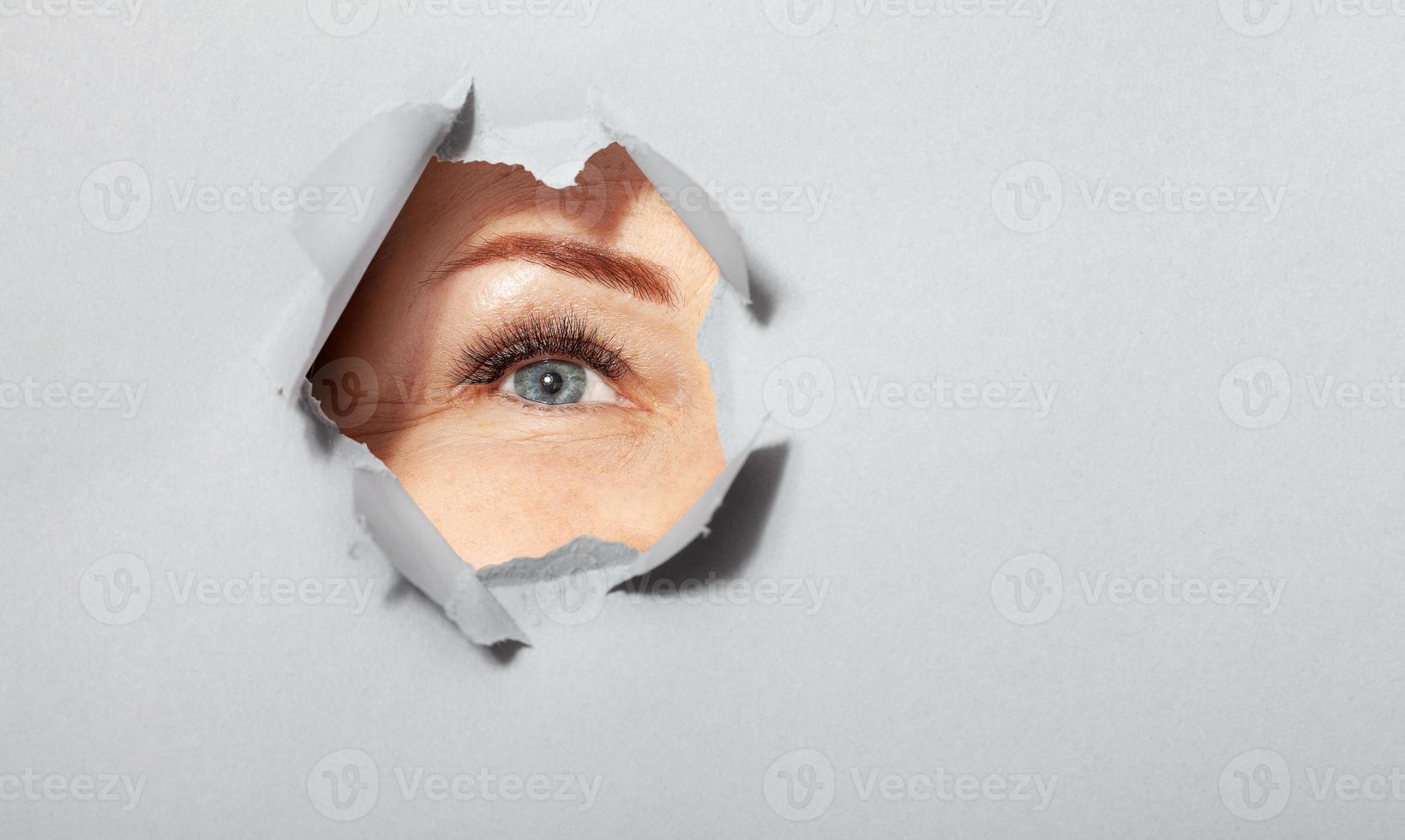 Part of the face one eye of a beautiful woman looks curiously through a torn gray paper background. Copy space. photo