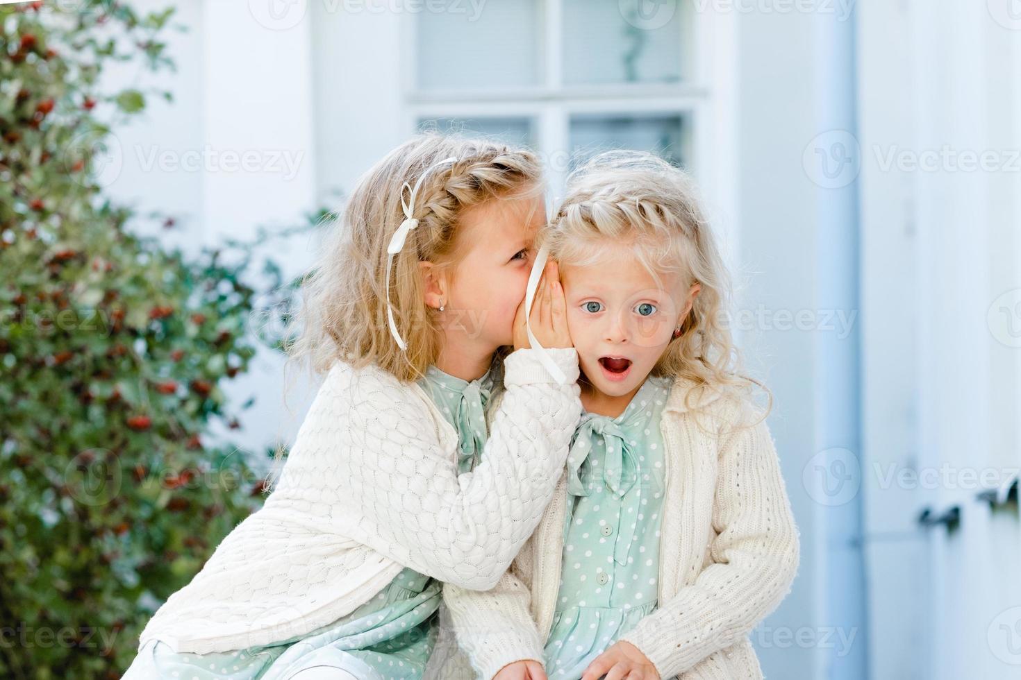 Two little cute girls are sitting in the garden and sharing secrets. One girl whispers something in another's ear. The two sisters have a close relationship. photo