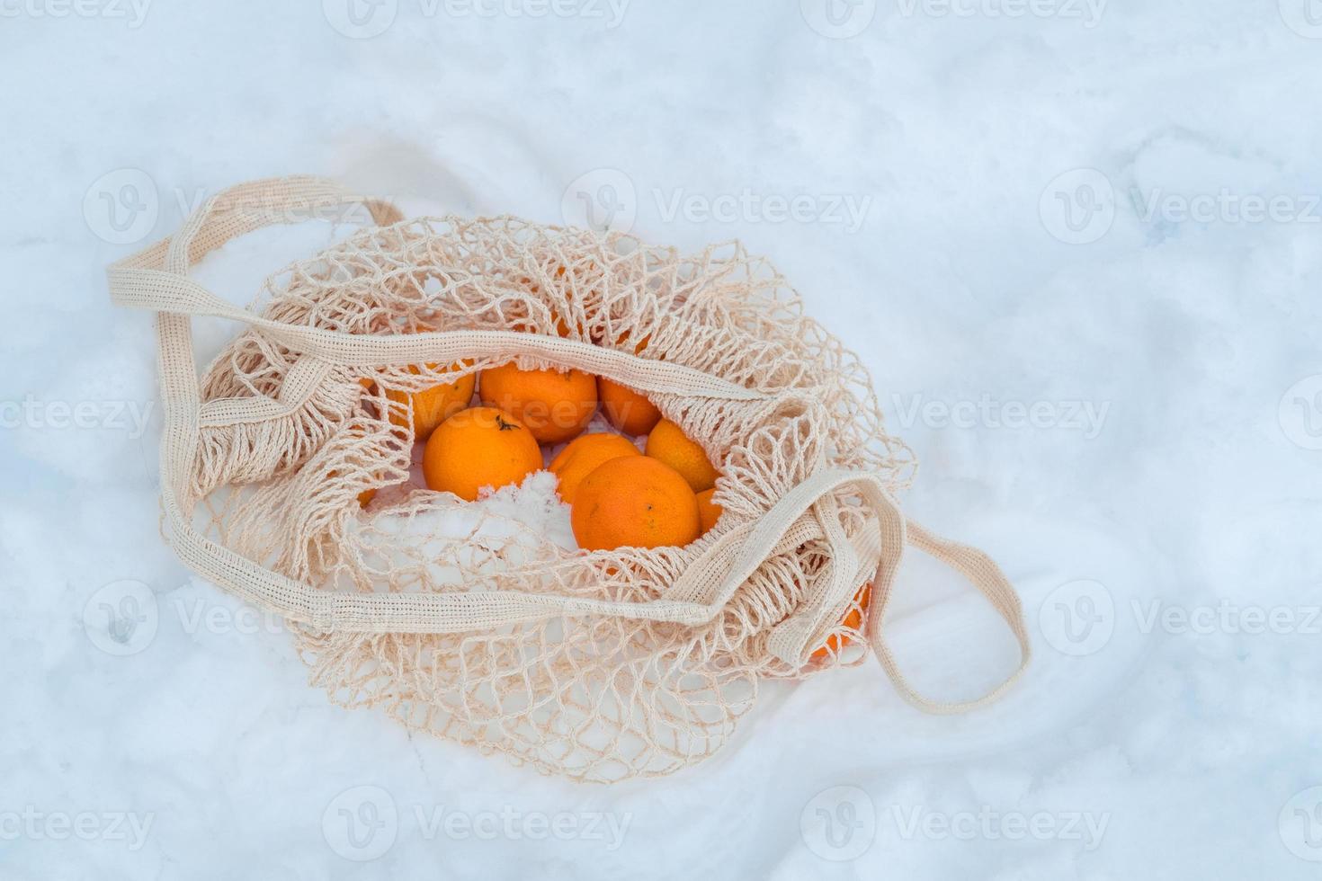 The tangerines are in a string bag, an organic mesh bag, on a bench that is covered in snow. Winter holidays concept photo