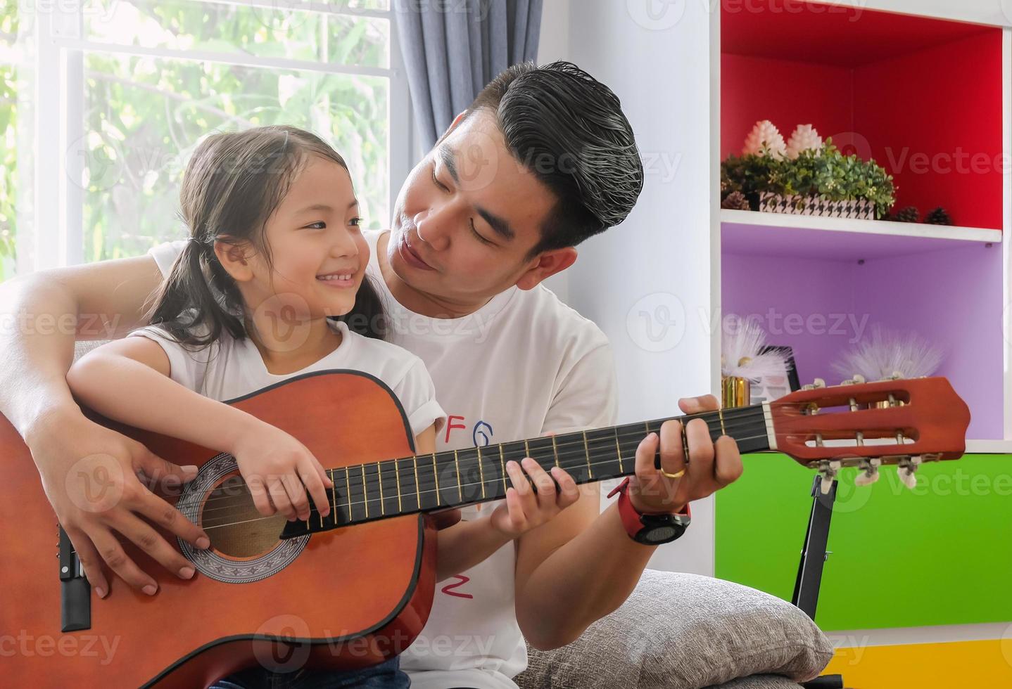 Young asian father and daughter family happy spending time together by playing guitar in living room warmth house background. Light effect and copy space. photo