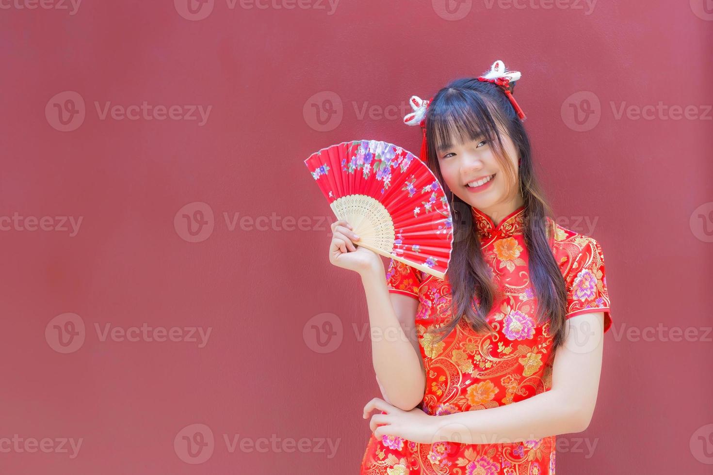 Beautiful Asian young woman in red cheongsam dress in Chinese new year theme stands smiling happily looking at the camera holds a fan on red background. photo