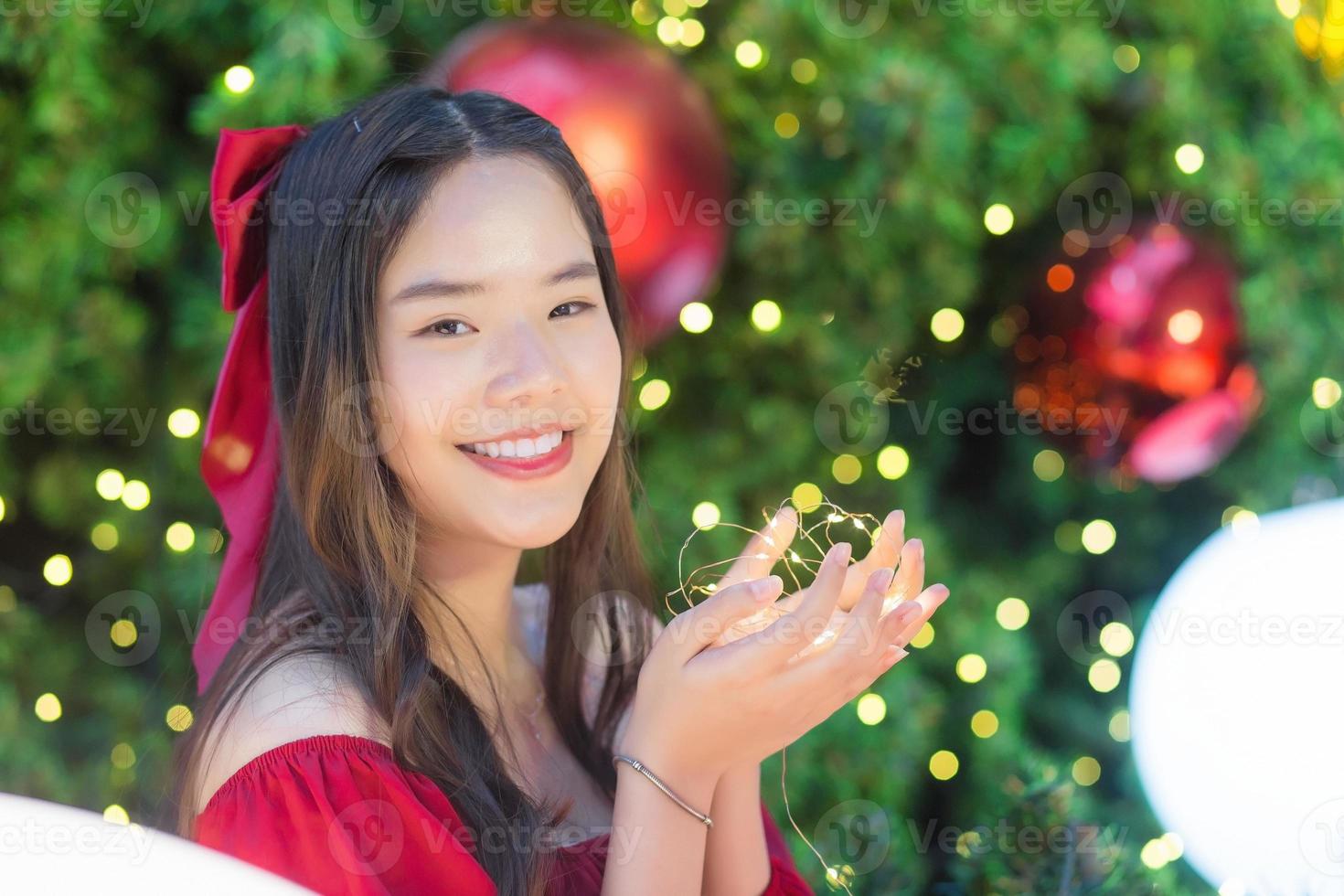 linda mujer asiática con vestido rojo se pone de pie felizmente. en su mano sostenía una luz frente al árbol de navidad. con bokeh como fondo en el tema de las celebraciones de navidad y año nuevo foto