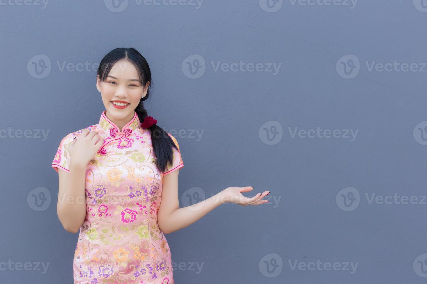 Beautiful Asian woman with long hair who wears pink Cheongsam dress in Chinese new year theme while her hand shows to present something and looks at camera smiling happily with the grey background. photo