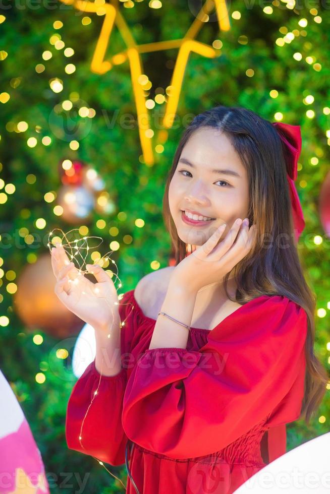 hermosa mujer asiática vestida de rojo se para felizmente en su mano, sostenía una luz frente al árbol de navidad con bokeh como fondo en el tema de las celebraciones de navidad y año nuevo foto