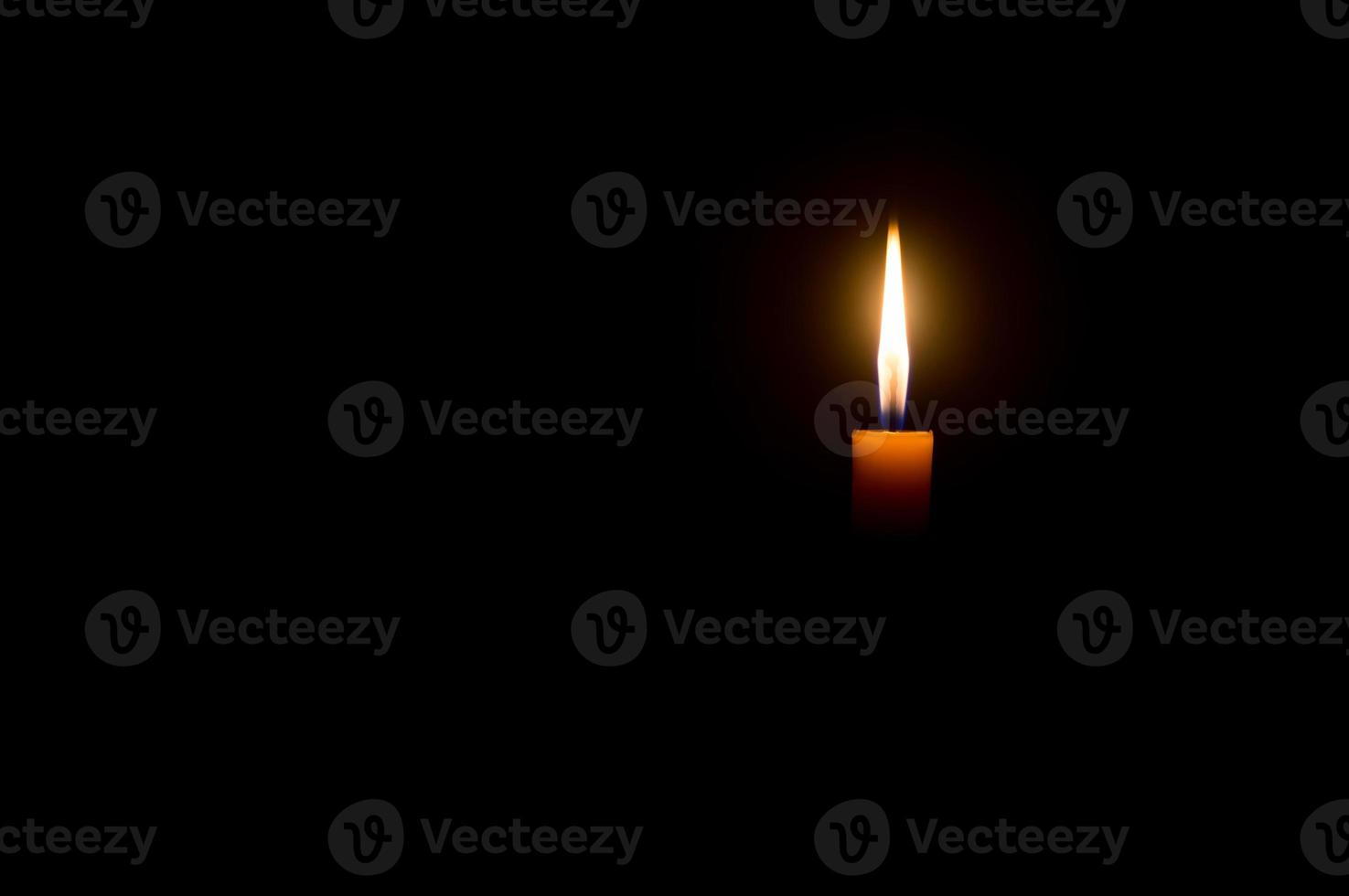 A single burning candle flame or light glowing on an orange candle on black or dark background on table in church for Christmas, funeral or memorial service with copy space photo