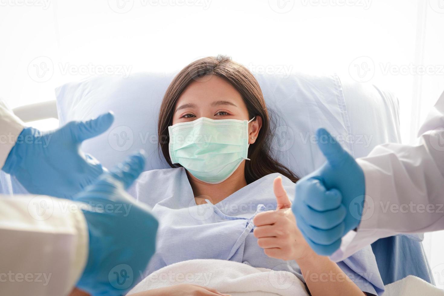una mujer asiática con una máscara está acostada en una cama de hospital. hay un grupo de médicos para tratar dar un pulgar hacia arriba para animar al paciente. el concepto de servicios médicos hospitalarios. foto