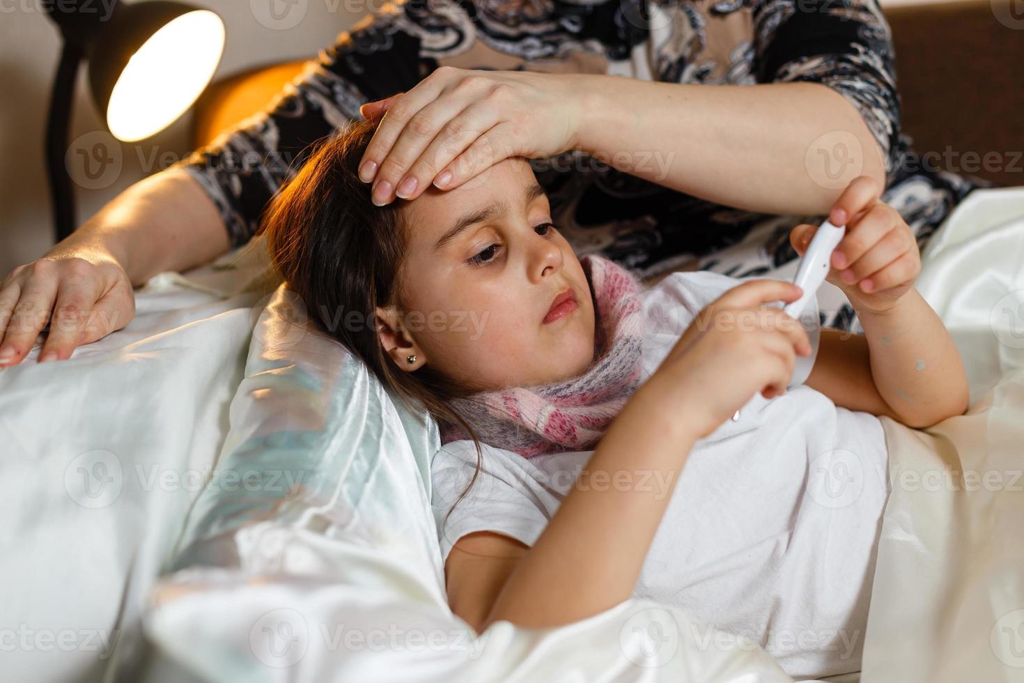 niña enferma en la cama. madre con termómetro está sentada cerca de la cama. foto