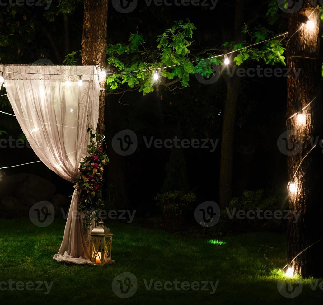 Wedding twilight ceremony decoration made of hanging on a tree golden lanterns and lamps and standing on the grass fired candles with the carpet in the middle photo