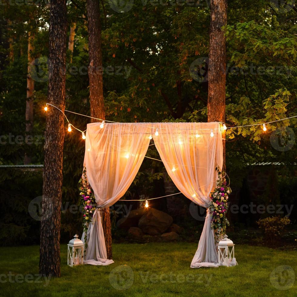 Wedding twilight ceremony decoration made of hanging on a tree golden lanterns and lamps and standing on the grass fired candles with the carpet in the middle photo