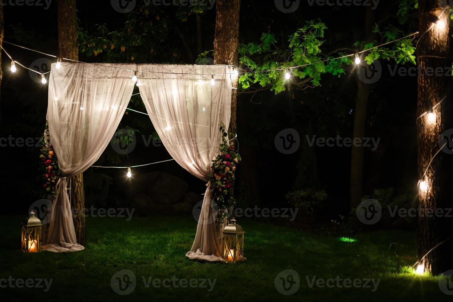 Wedding twilight ceremony decoration made of hanging on a tree golden lanterns and lamps and standing on the grass fired candles with the carpet in the middle photo