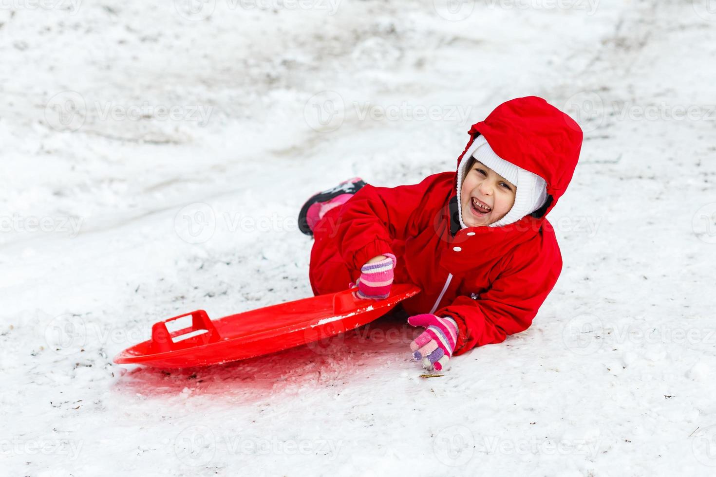 una niña muy sonriente con su traje de esquí deslizándose por una pequeña colina cubierta de nieve con su trineo foto