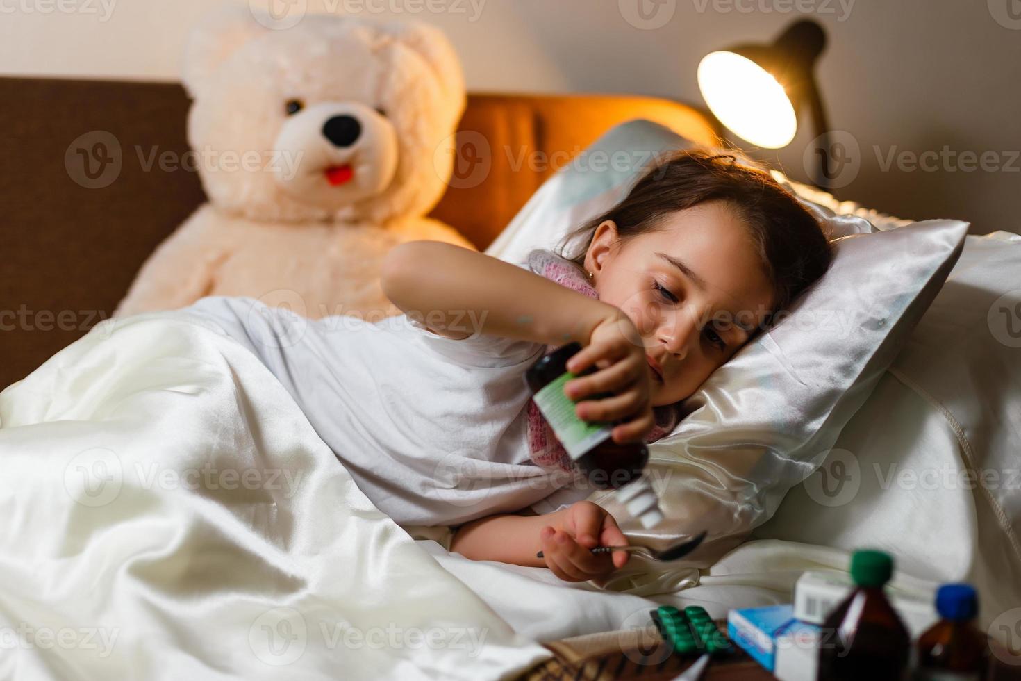 la niña enferma cubierta con una manta está abrazando al oso de peluche y mirando tristemente la medicina mientras está acostada foto