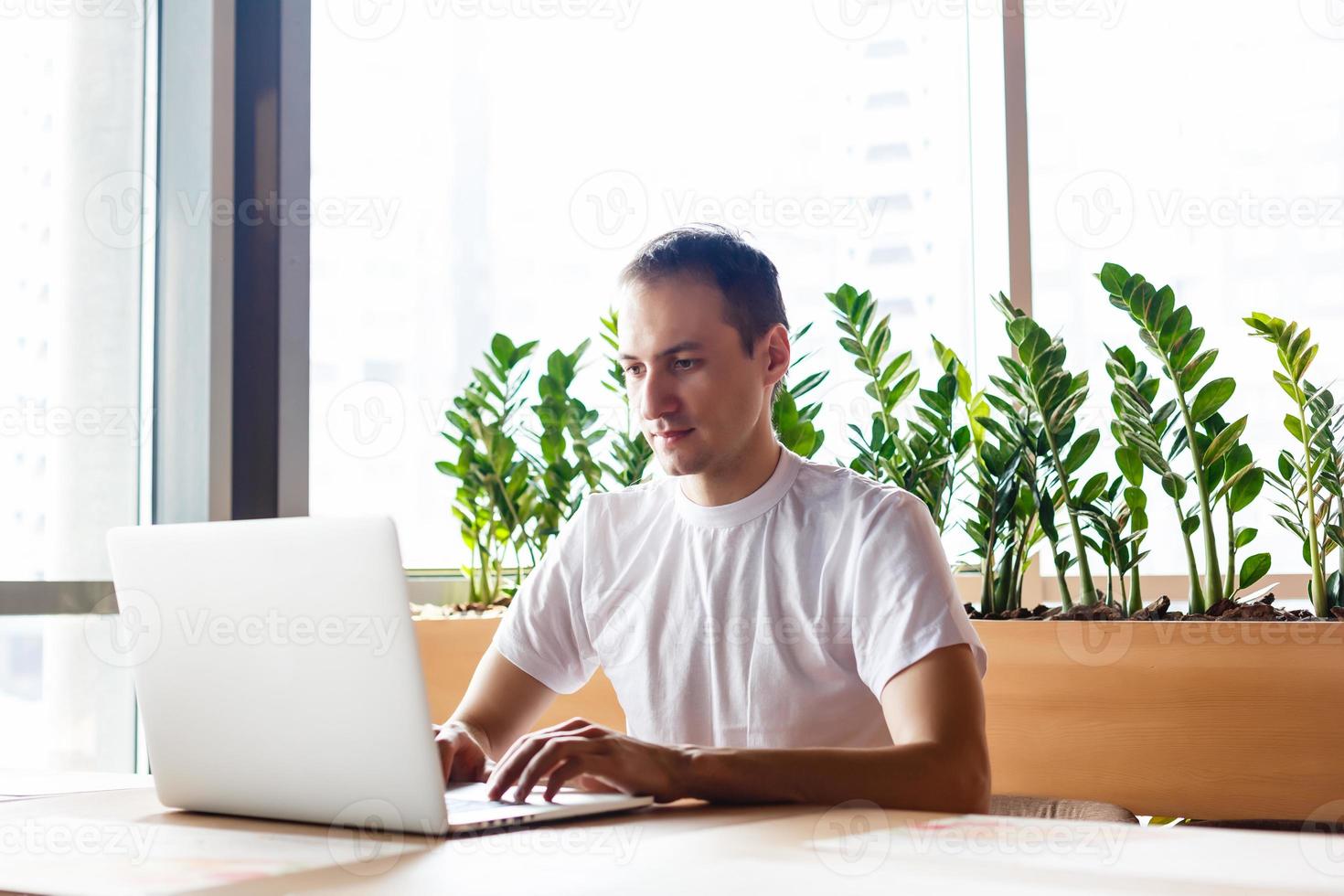 Young businessman holding mug of coffee while working on laptop computer in store with healthy food, male freelancer writing text on laptop keyboard while enjoying cup of cappuccino in modern cafe photo