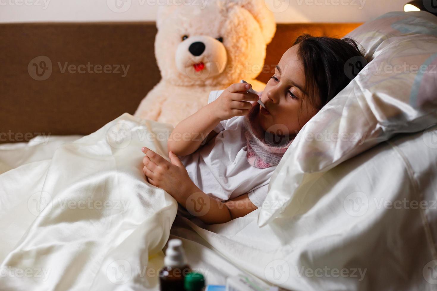 Sick girl with a thermometer in mouth lying in bed grumpy face photo
