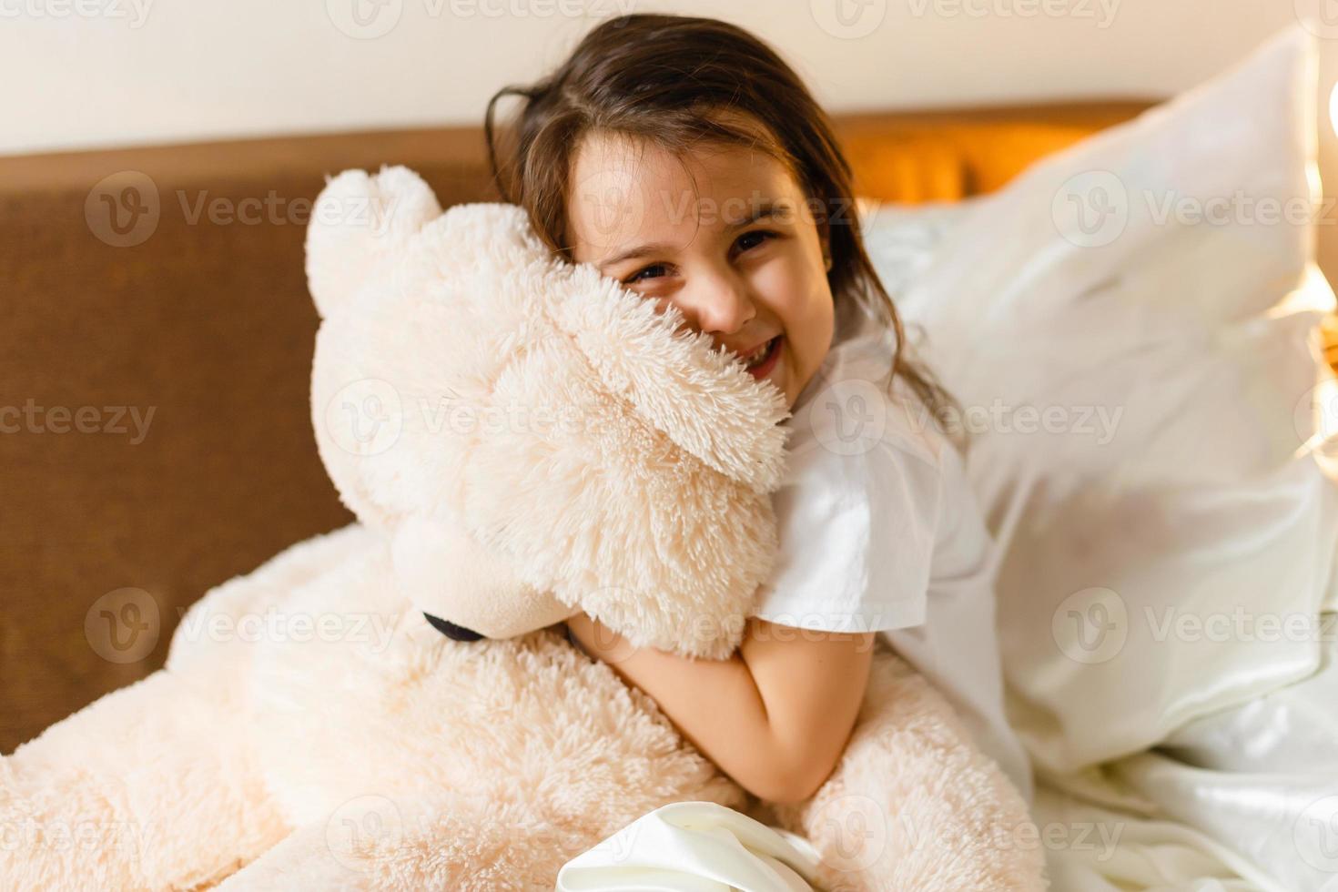 Sweet little girl is hugging a teddy bear looking at camera and smiling while sitting on her bed 396815581 photo