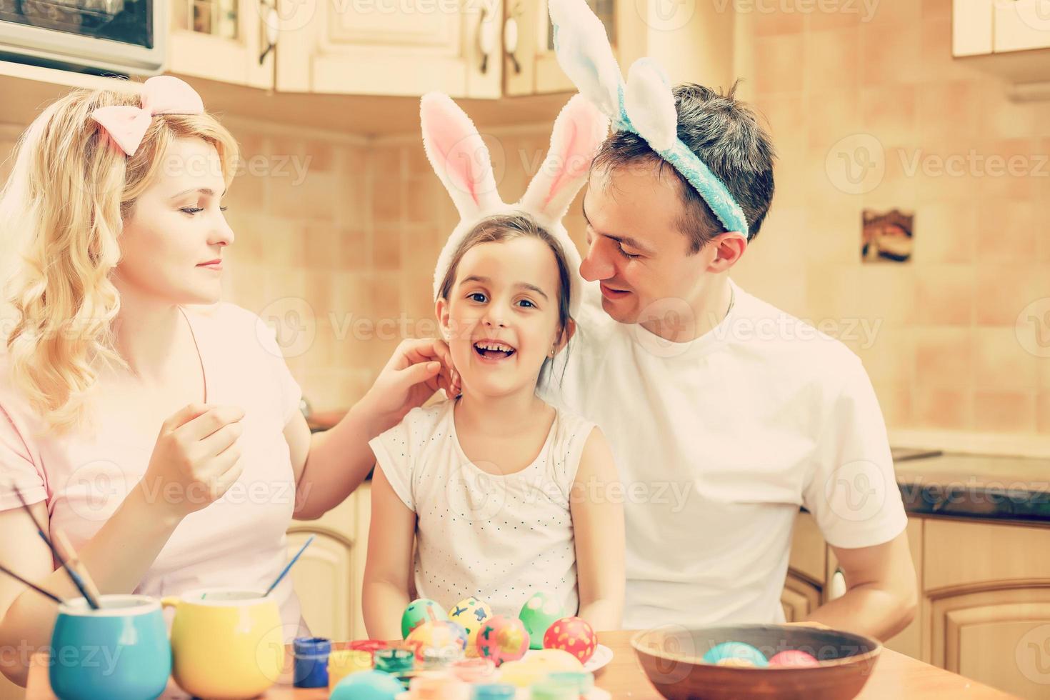 Mother, father and daughter are painting eggs. Happy family are preparing for Easter. Cute little girl wearing bunny ears. photo