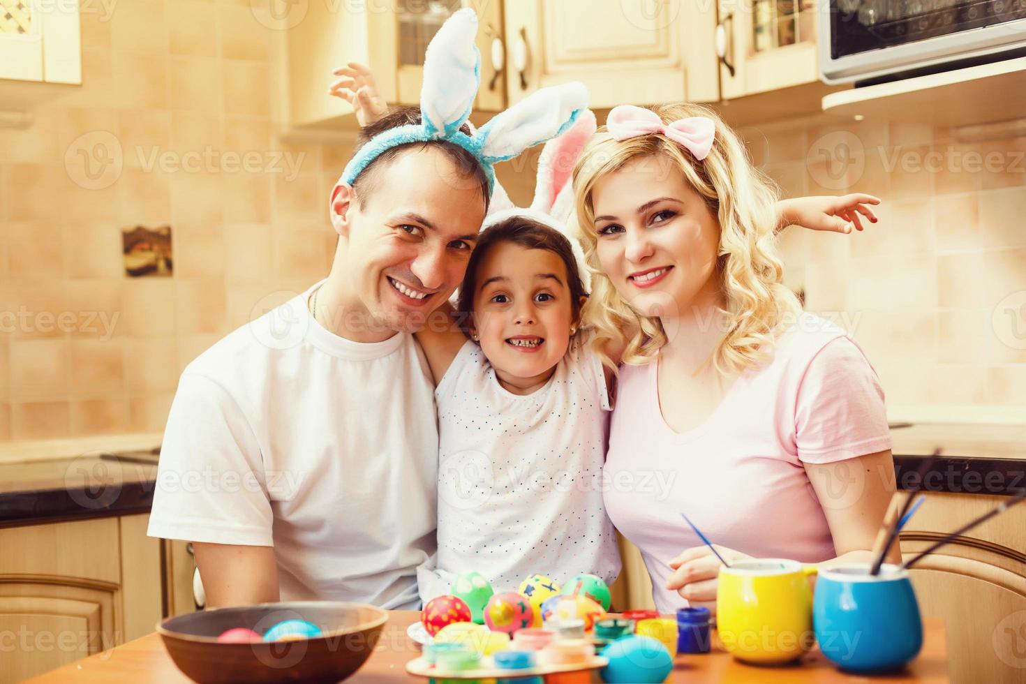 Mother, father and daughter are painting eggs. Happy family are preparing for Easter. Cute little girl wearing bunny ears. photo