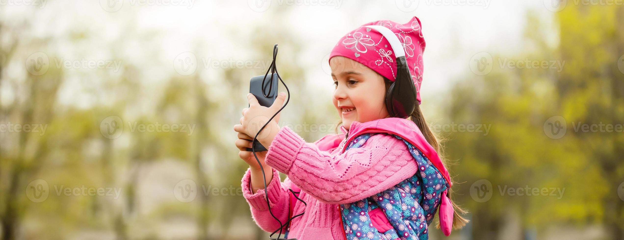 Side view of happy young girl child wearing colorful headphones, listening to music on the smartphone outdoors in summer. photo