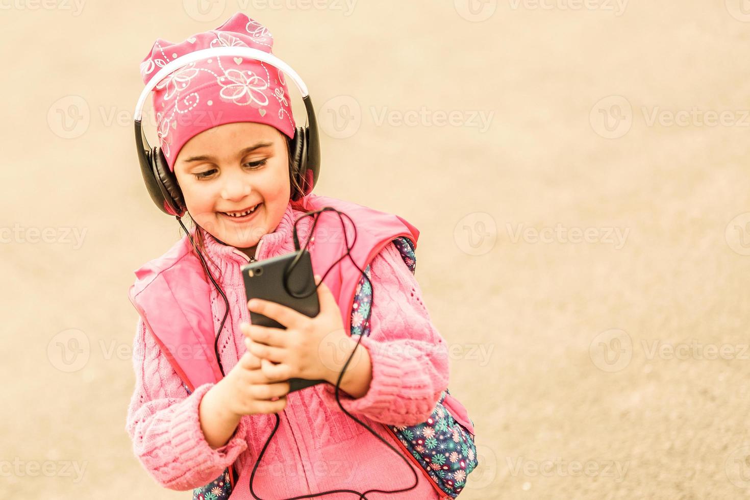 Side view of happy young girl child wearing colorful headphones, listening to music on the smartphone outdoors in summer. photo