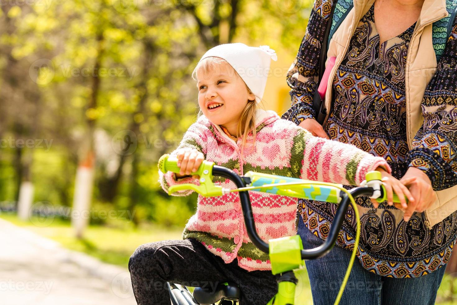 Beautiful and happy young mother teaching her daughter to ride a bicycle. Both smiling and looking at each other. Summer park in background. photo
