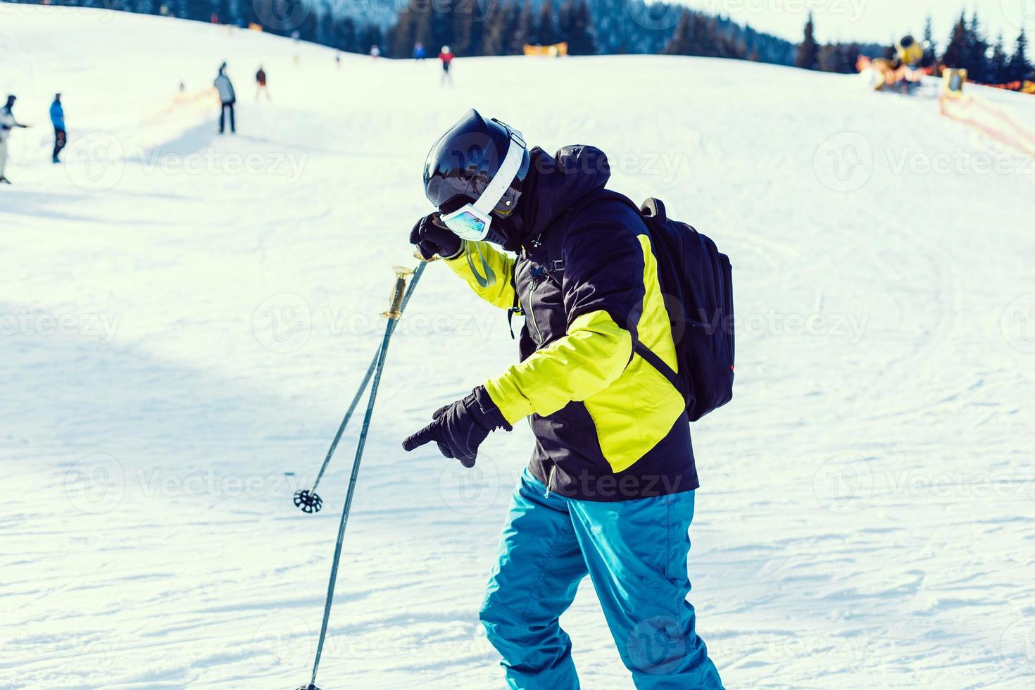 Skier holding ski and looking at beautiful snow covered mountains. Young man thinking and looking snowy mountain with copy space. Side view of winter skier guy with mask holding ski equipment outdoor. photo