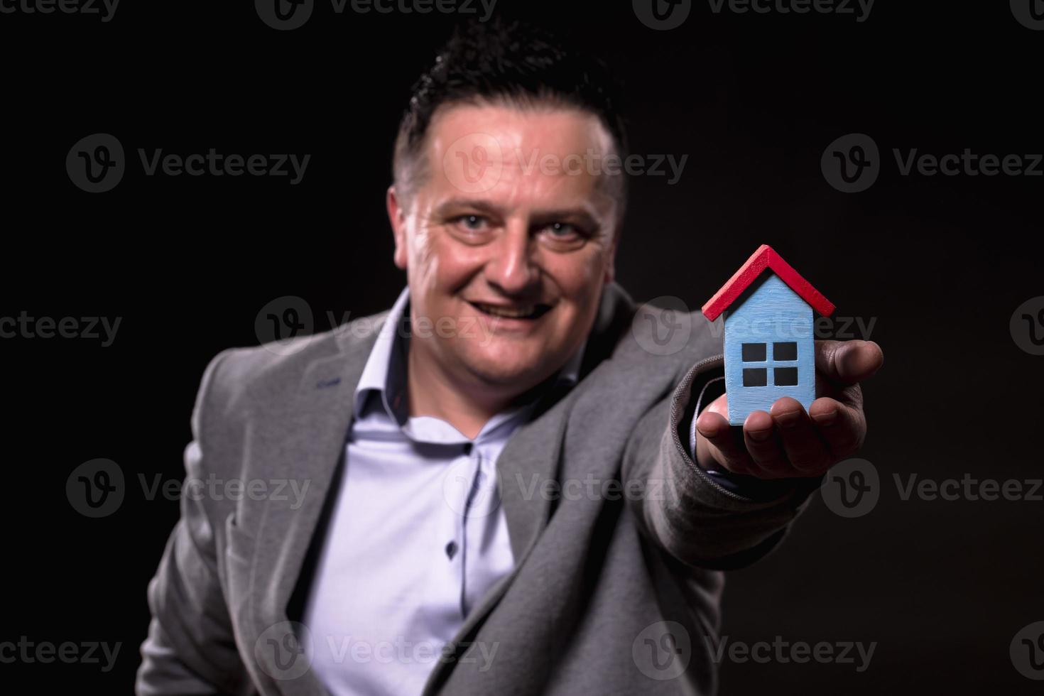 retrato de un hombre emocionado vestido con ropa formal sosteniendo una casa en miniatura contra un fondo negro. concepto de compra de casa. foto