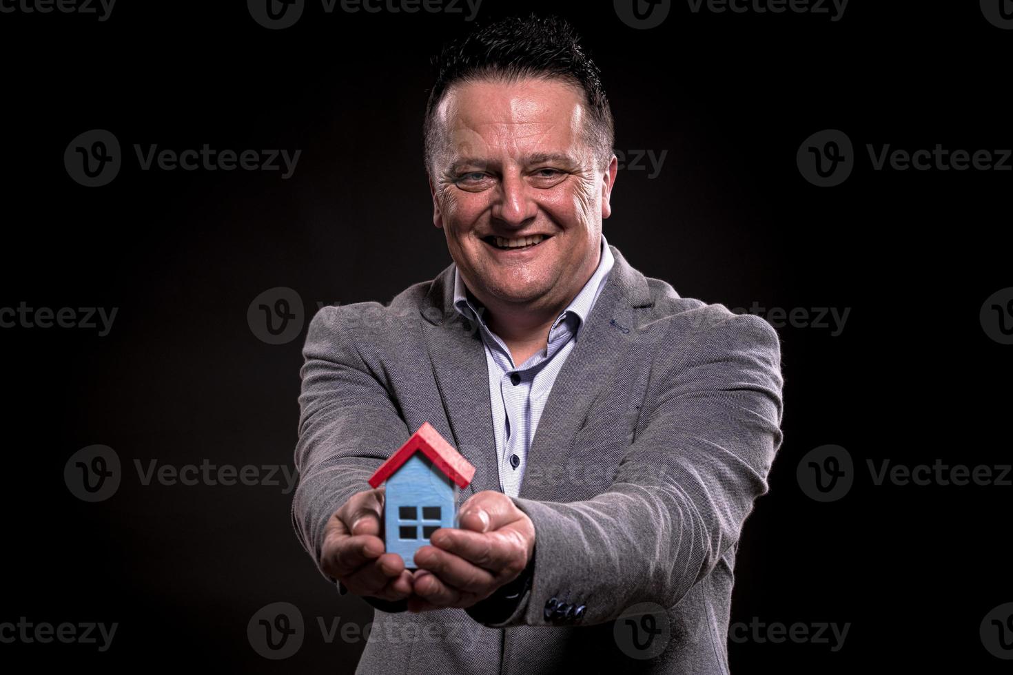 retrato de un hombre emocionado vestido con ropa formal sosteniendo una casa en miniatura contra un fondo negro. concepto de compra de casa. foto