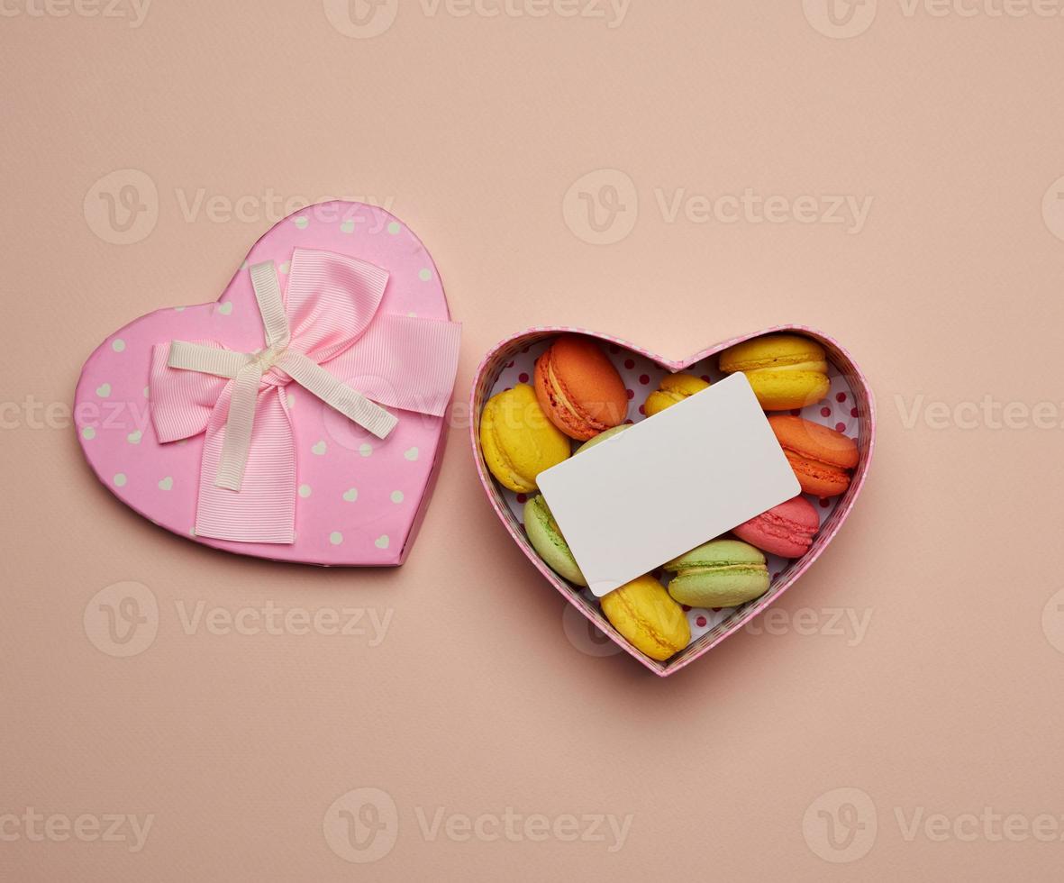 round baked multicolored macarons lie in a pink cardboard box in the shape of a heart on a beige background photo