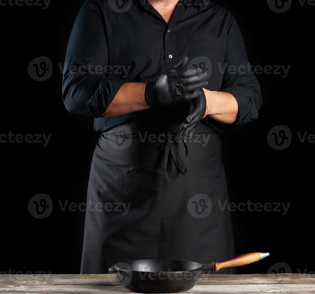 el chef con camisa negra y delantal se pone guantes de látex negros en las manos antes de preparar la comida foto