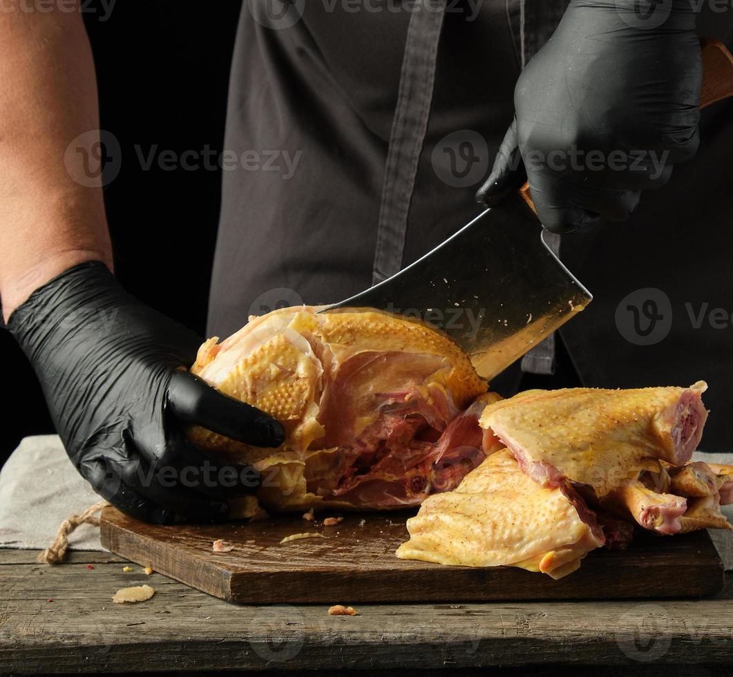 chef in black uniform and latex gloves chopping raw chicken into pieces on a brown wooden board photo