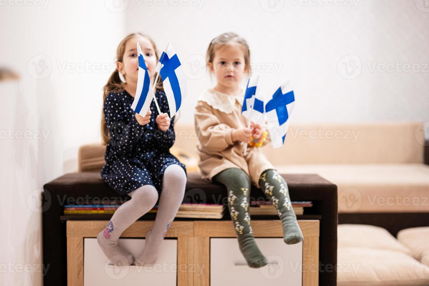Two sisters are sitting on a couch at home with finnish flags on hands. Finland children girls with flag . photo