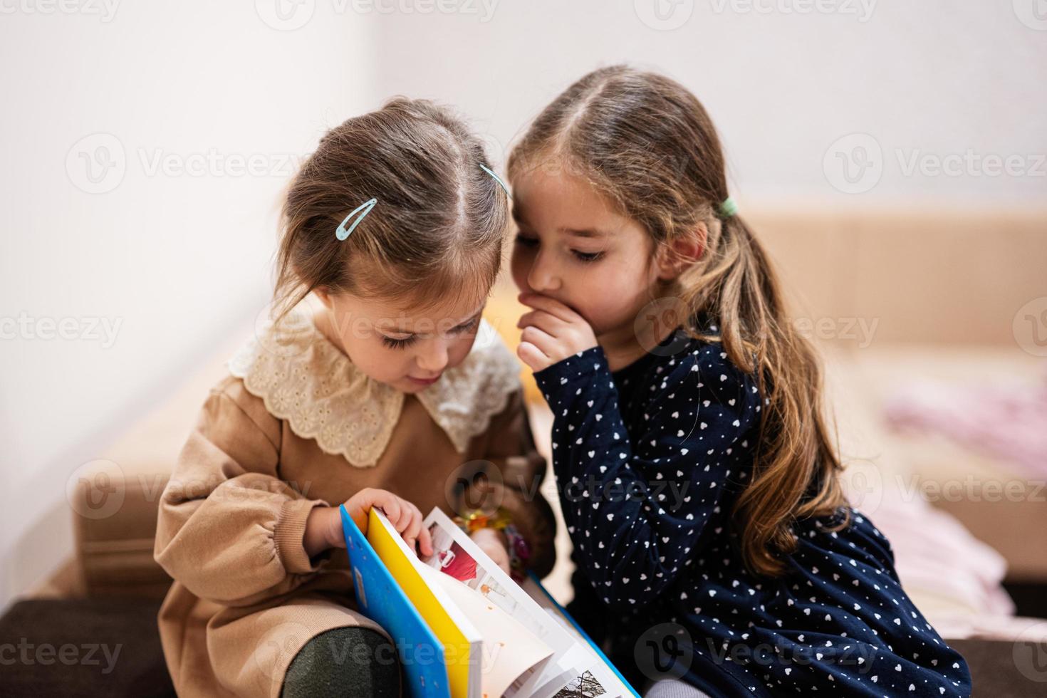 Two sisters are sitting on a couch, looking and read a book together. photo