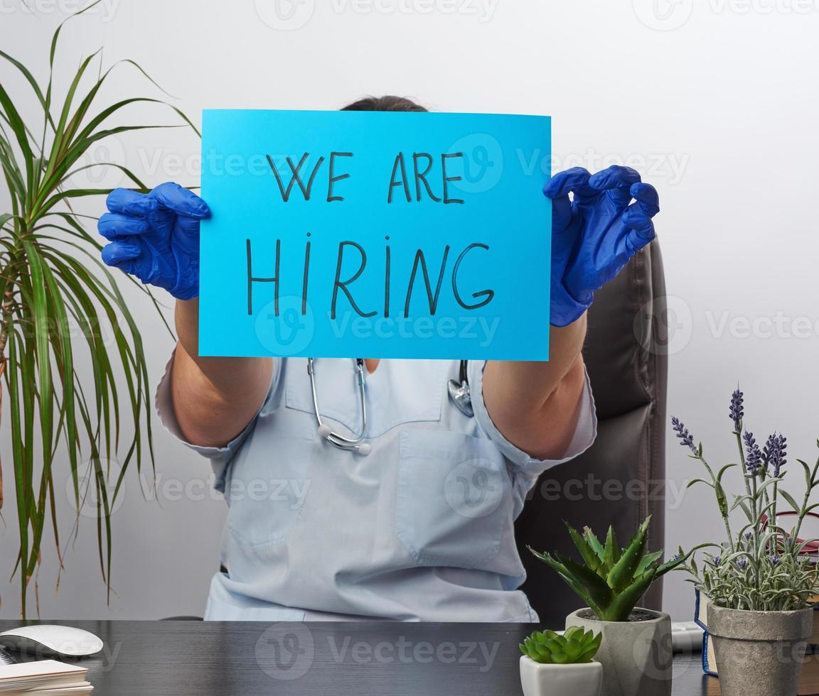 doctor in a blue coat, sterile medical gloves holds a poster with the inscription we are hiring photo