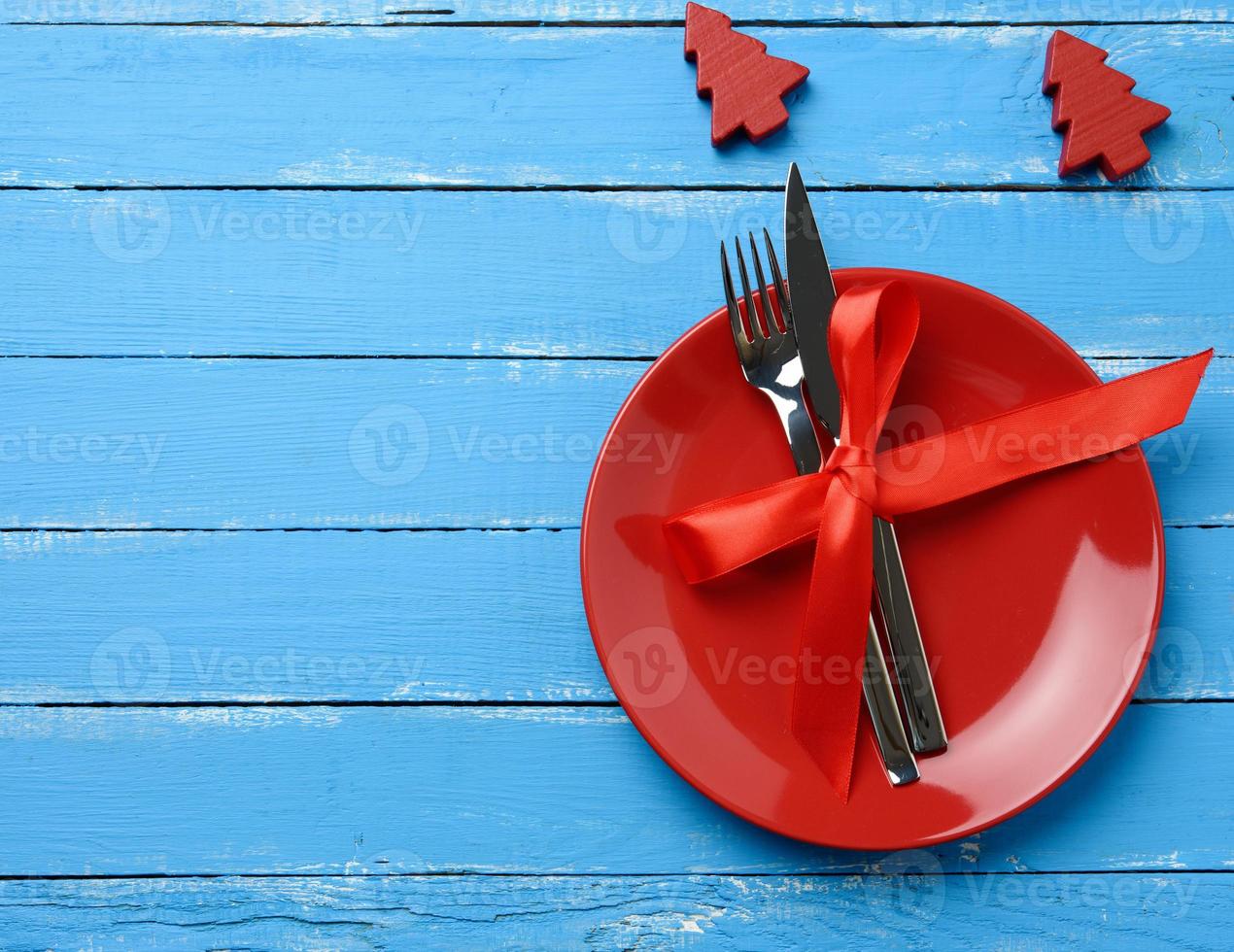 round plate and metal fork and knife tied with a red silk ribbon, blue wooden background photo