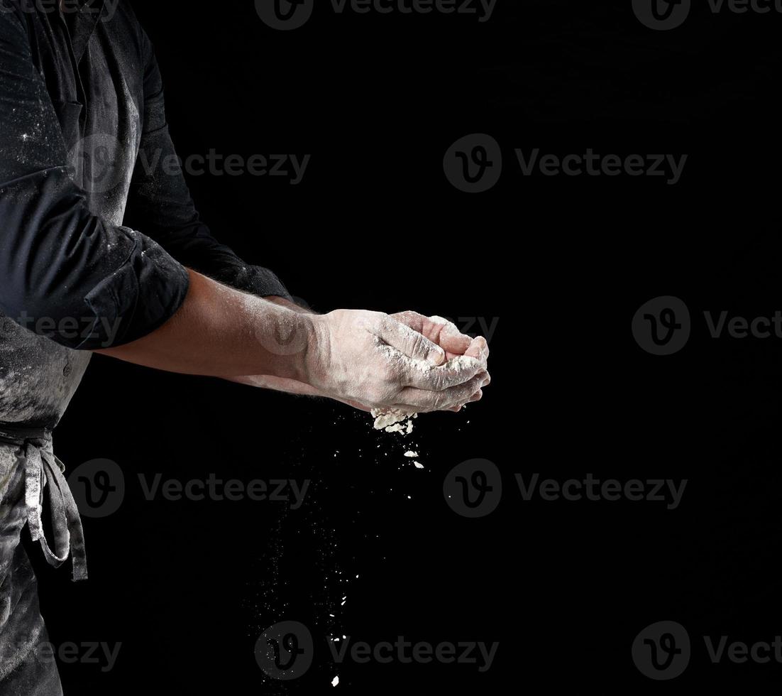 baker in black clothes holds white wheat flour in palms on black background photo