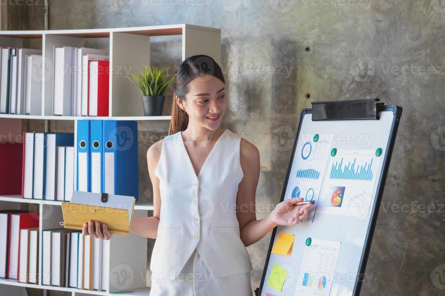 Asian businesswoman presenting business data with a board with charts, graphs hand holding a yellow notebook, And business documents lying on the table in the office, business concept. photo