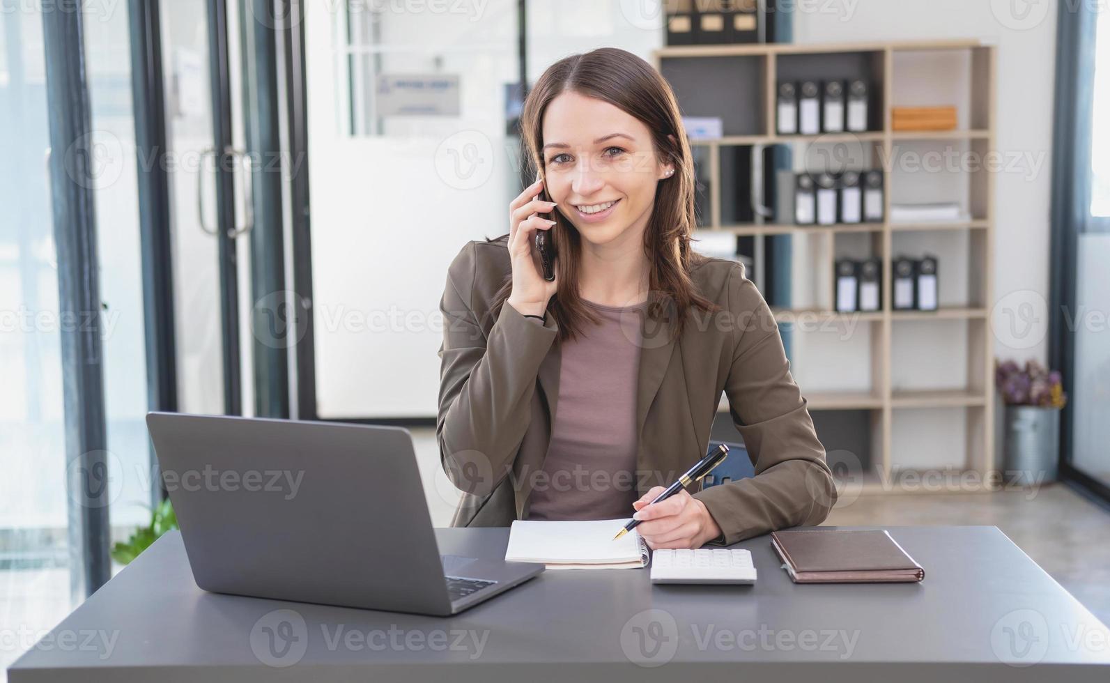 Marketing, Finance, Accounting, Planning, Businesswoman of bi nationality is talking with a customer representing a company distributor using a smartphone with laptop pen and notepad on office desk. photo