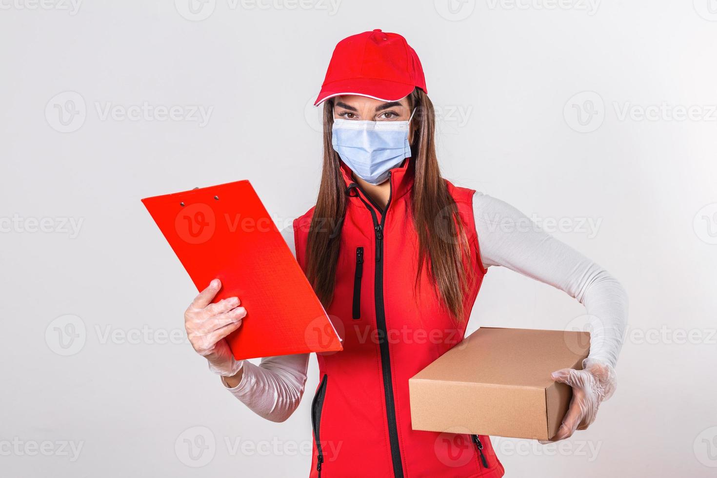 repartidor entregando paquetes con portapapeles y paquete sonriendo feliz en uniforme rojo. Hermosa mujer joven con máscara médica y guantes mensajero profesional aislado sobre fondo blanco. foto